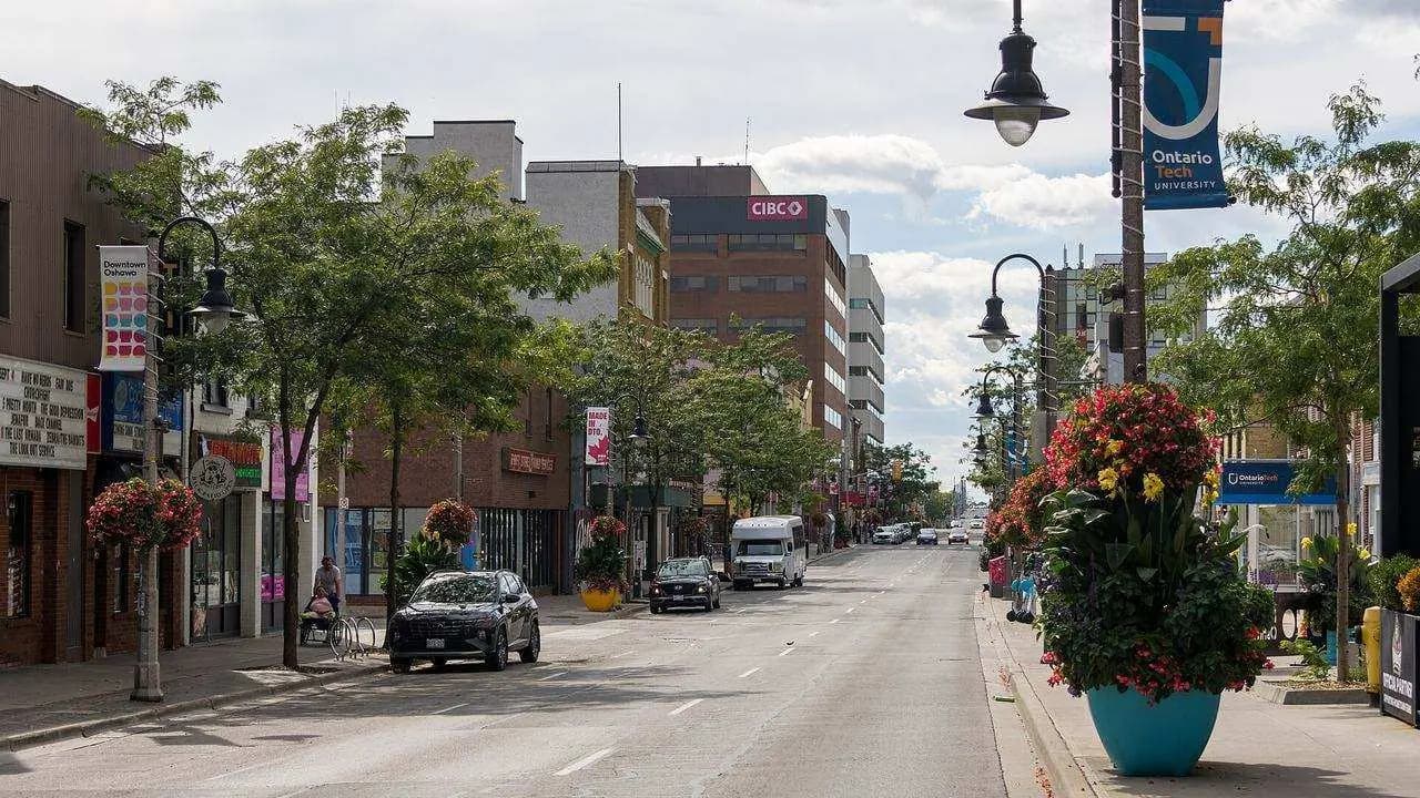 Looking west along King Street in downtown Oshawa, with storefronts, trees and flower planters; two distant people on the sidewalk are not identifiable.