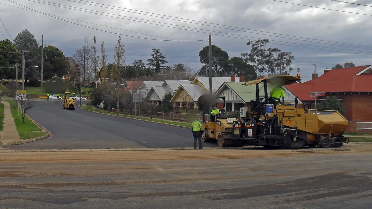 A road being resurfaced with a roller and paving machine at work; workers in high-visibility clothing are distant with faces blurred and not identifiable.