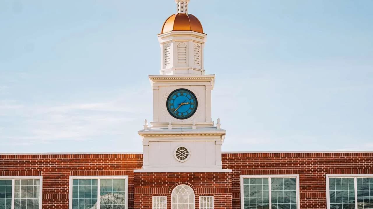 Brick school building with a white clock tower and copper dome, seen across an empty paved forecourt.