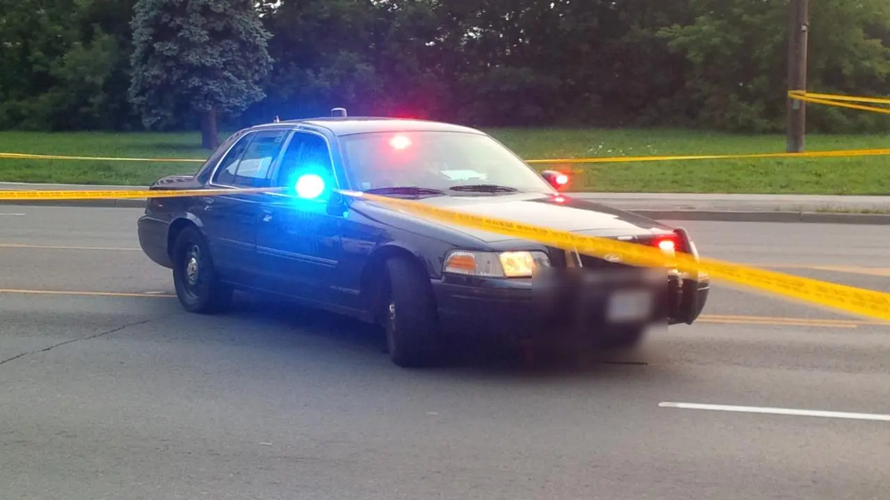 A dark unmarked police sedan with red and blue lights flashing behind yellow police tape on a road; no people visible.